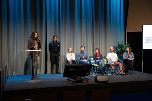 Kuvassa lava, jossa yksi tutkija seisoo ja puhuu mikrofoniin ja muut istuvat rivissä.