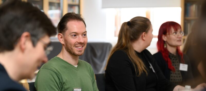 Neljä henkeä pöydän ääressä. Vihreäpaitainen mies keskellä hymyilee.