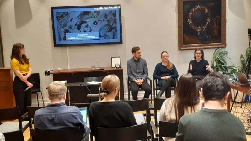 Kuvassa istumassa kolme panelistia ja seisomassa puheenjohtaja sekä yleisön edustajien selkiä.