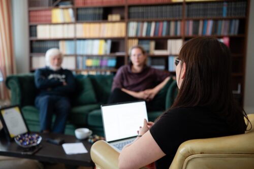 Kuvassa haastattelija istuu selin kameraan. Vihreällä sohvalla istuu kaksi haastateltavaa.